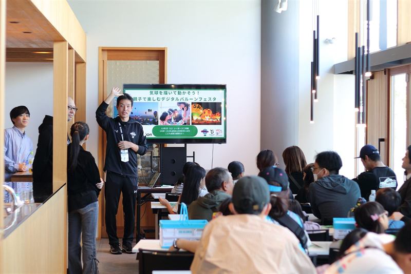 会場でイベントの進め方を説明するスタッフと着席した参加者